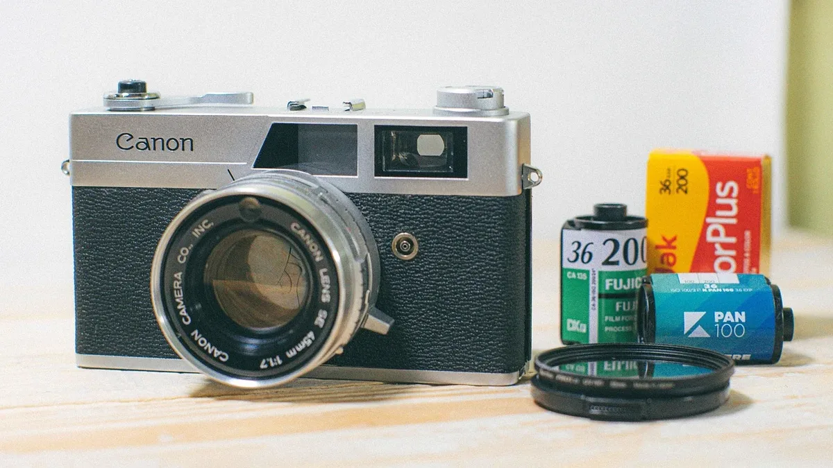 A Canon camera with a lens attached, accompanied by a roll of film and a few other camera-related items on a light-colored surface.